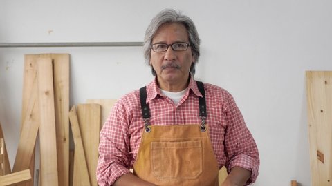 Portrait of a happy senior Asian man wearing a lumberjack shirt and apron, smiling, crossed arms, showing thumbs up, in the wood workshop. Front view, indoors, medium shot, carpenter and DIY concept