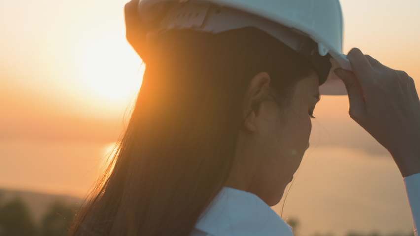A woman engineer is putting a protective helmet on her head at sunset.
