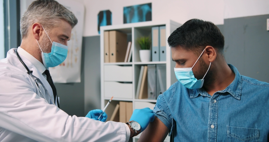 Close up portrait of experienced Caucasian middle-aged male specialist at work in quarantine doing vaccine to young Hindu man patient in medical mask in hospital, vaccination, coronavirus treatment | Shutterstock HD Video #1066881712