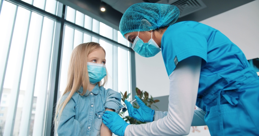 Close up of professional healthcare specialist female nurse in protective uniform and medical mask making covid-19 vaccine injection to little adorable kid girl in clinic lab room, vaccination concept | Shutterstock HD Video #1066881853