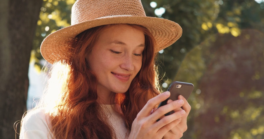 Young Woman in Straw Hat typing message on Mobile Phone in park and smiling. Happy Student Taking a Rest on the Weekend. Having curly Ginger Hair. Looking attractive and with Good Mood.