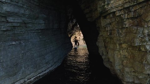 Young athletic man rowing with paddle standing on SUP board and swim into narrow crevice between cliffs, pushes off with an oar from the rocks, looks around and admires the beautiful view around.