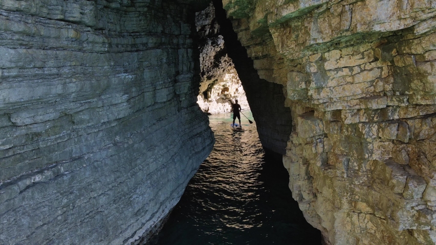 Young athletic man rowing with paddle standing on SUP board and swim into narrow crevice between cliffs. Active recreation and healthy lifestyle.
