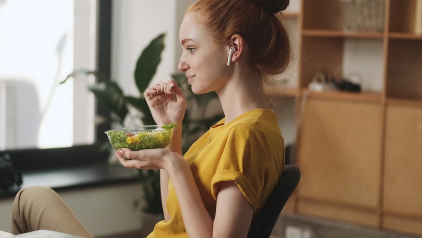 A focused young redhead woman with earbuds is eating salad while watching laptop sitting in the office