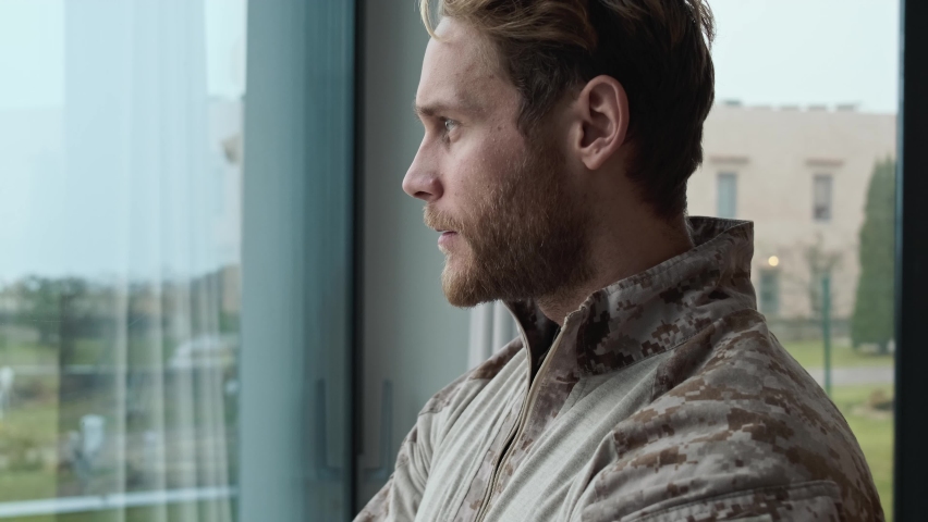 A focused young american soldier man is looking to the window standing inside