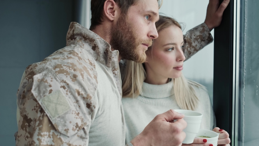 A calm couple young american soldier man and blonde woman are drinking tea while looking to the window standing inside the apartments