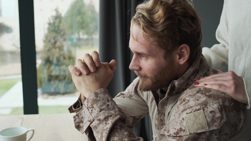 A side view of a calm soldier man is looking to the window sitting at the table while his girl friend is massaging his shoulders inside the apartments