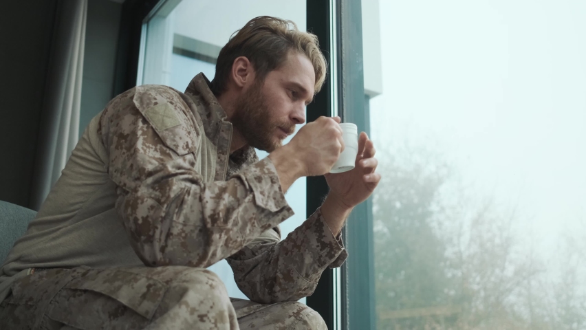 A handsome young american soldier man is looking to the window sitting inside the apartments and drinking tea while his woman is coming and hugging him