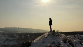  Silhouette of a person standing on a top. Man on snowy peak, drone fly around man. In background town and bridge. Aerial footage man on top in winter time. Can use in motivation video.  