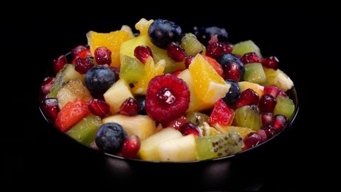 Colorful fruit salad in a bowl rotating on a black background. Close up of fresh and tasty salad with orange, apple, berries, pomegranate, kiwi, ananas, raspberries, blueberries and strawberries.