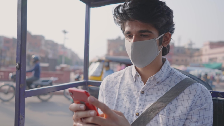 close shot of a young attractive Indian male office employee with a protective face mask sitting in a moving local auto-rickshaw using a mobile phone to type a text message amid the COVID 19 epidemic Royalty-Free Stock Footage #1066900849