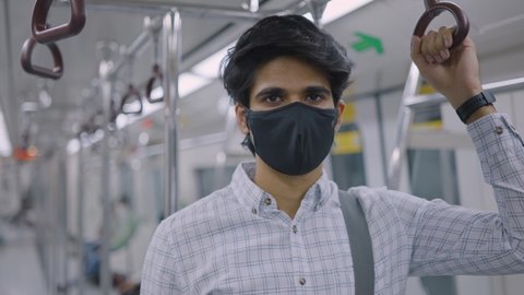 Shot of young Indian attractive male corporate office employee wearing protective face mask traveling to workplace standing in a moving city metro train looking at the camera amid COVID 19 epidemic