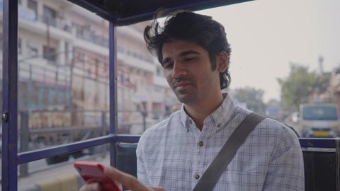 close shot of a young attractive Indian male office employee is smiling and using a mobile phone to type a text message in a moving an auto-rickshaw through a busy city market road  