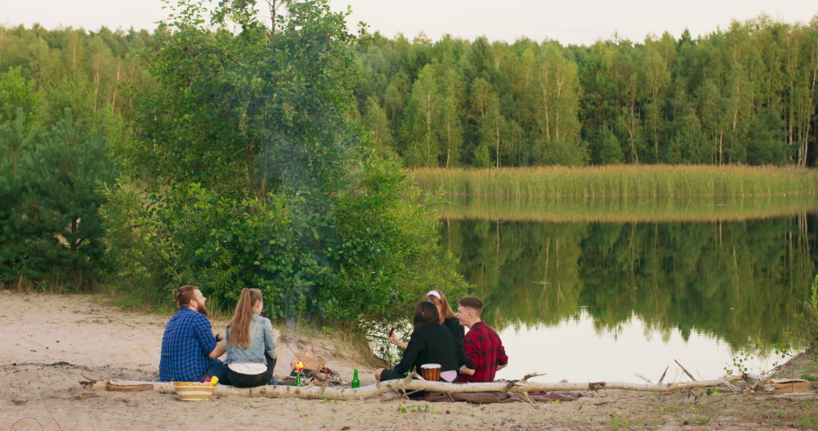 Friends sit around bonfire talking and drinking beer on sandy beach. Young mixed race group of men and women with beverage singalong