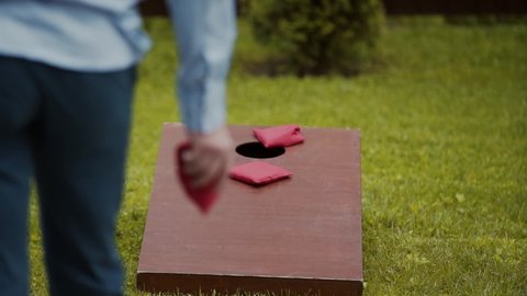 Cornhole. A man in blue trousers and a shirt throws red bags of corn, the bags do not hit the target, fly past a wooden hole