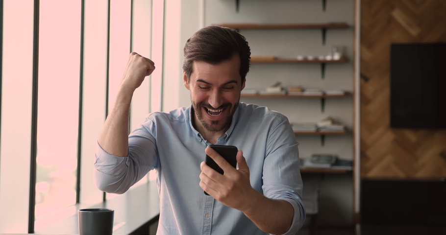 Overjoyed euphoric young caucasian man reading message on cellphone with unbelievable news, celebrating mobile game victory, winning lottery or gambling, feeling amazed in modern office room. | Shutterstock HD Video #1066905025