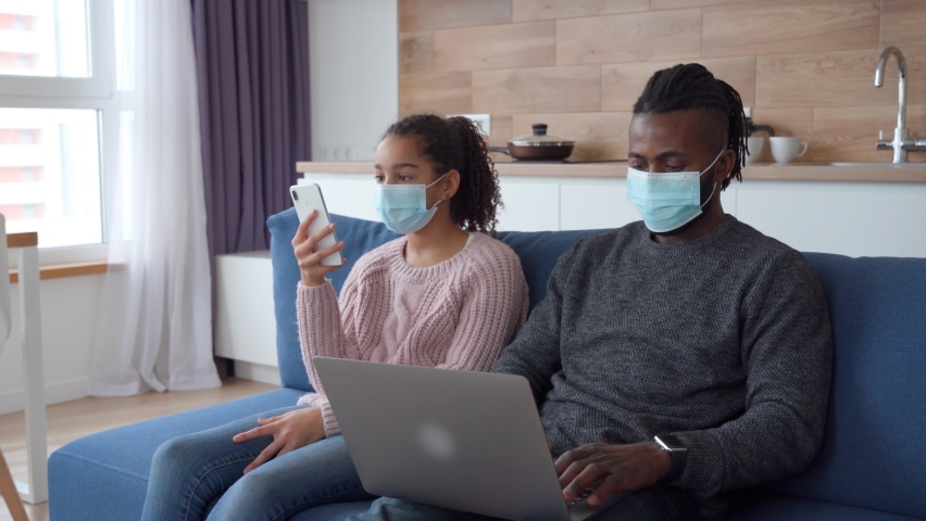 Lifestyle portrait of Afro American family sitting at home in protective face masks and using gadgets. Young father use laptop and his teen age nice daughter shows him app on smartphone in quarantine.