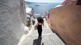 Tourist travel woman in Oia, Santorini, Greece. Happy elegant fashion model in black walking on stairs by famous landmark destination in Greece Santorini