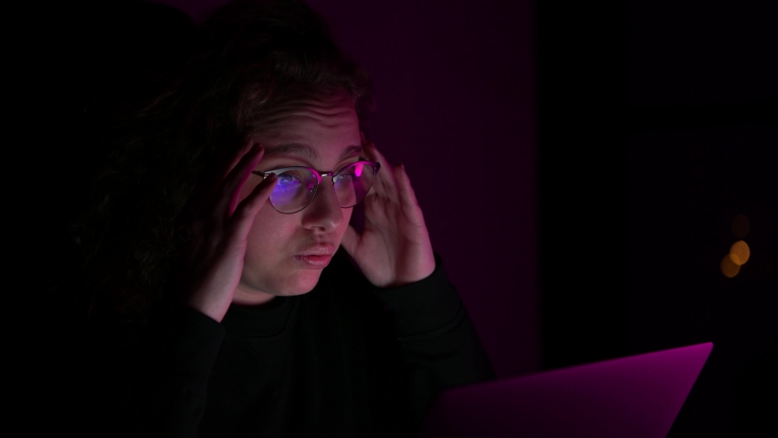 4K, An overwhelmed woman raises her hands to her head while she is browsing on her computer. It is night and it is dark. There are lights of different colors. | Shutterstock HD Video #1066912201