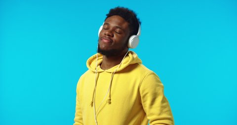 Close up portrait of happy relaxed young handsome African American man dancing rhythmically moving listening to music in headphones posing isolated on blue background in studio enjoying sounds