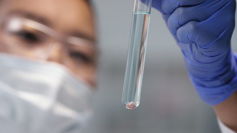 Close-up of female Asian scientist in safety mask and glasses looking at test tube with water and tiny piece of artificial cultured meat