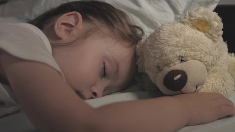 The kid sleeps at home on the sofa in the children's room. The sleeping baby is happy and carefree in bed, hugging a teddy bear toy. The mother covered her child with a blanket. Happiness in a dream.