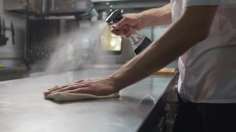 Slow-motion close up of unrecognizable chef cleaning his workplace spraying sanitizer over kitchen table surface wiping it with cloth