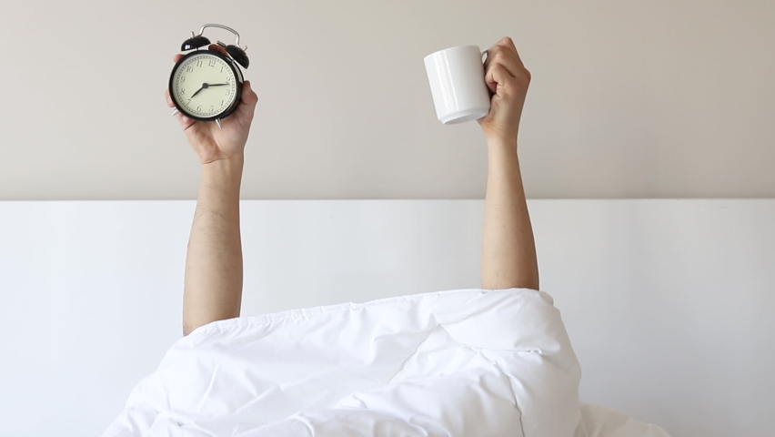 Man showing arm raised up holding coffee cup and black alarm clock behind duvet in the bed room, Young boy with two hands sticking out from the blanket. wake up with fun in morning concept. | Shutterstock HD Video #1066932136