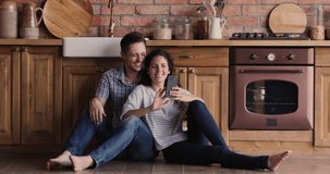 Full length happy positive caucasian married couple resting on warm floor in kitchen, choosing discussing goods in internet store, shopping online using modern technology cellphone gadget indoors.