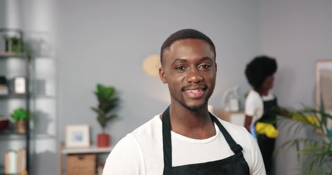 Close up portrait of joyful African American handsome young male worker in apron standing in room looking at camera and smiling, professional cleaner, cleaning service industry, work concept