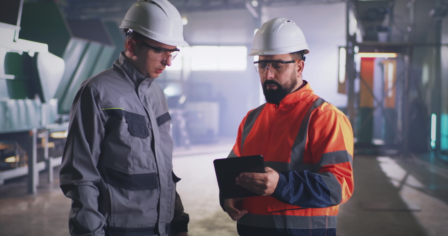 Male employee in uniform and hardhat looking around and showing factory workshop to inspector with tablet | Shutterstock HD Video #1066937083