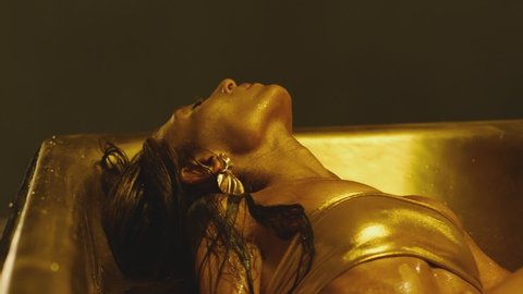 Footage of stylish woman on bikini and with shining glitters in her beautiful body swimming and moving inside bathtub . Girl lying in water in bathroom interior with golden or yellow color . Close up