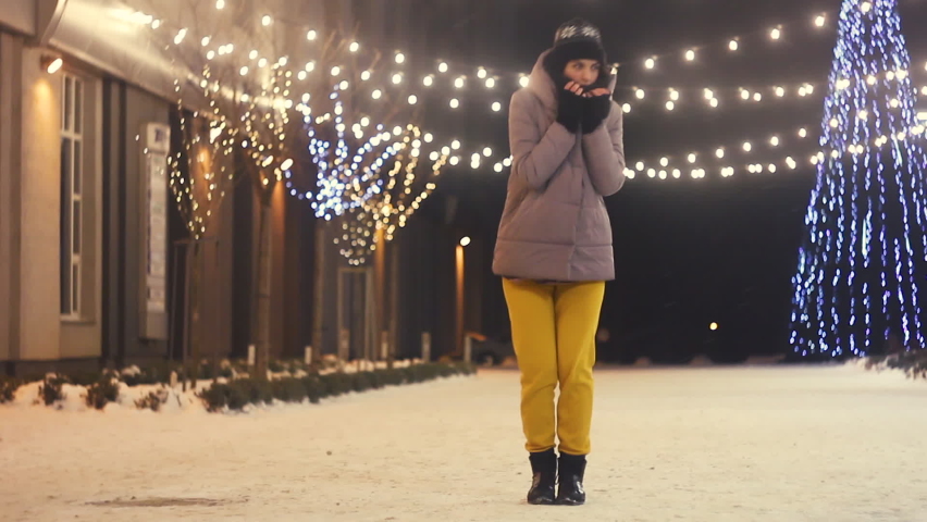 Young woman in yellow pants freezes on the street, trying to warm up, standing under the christmas lights