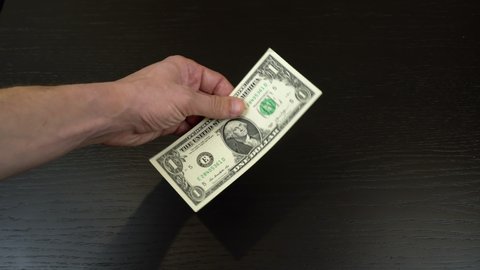 A man's hand on a black background holds one American dollar in his hand between his fingers, and twists and defiantly shows it from all sides.