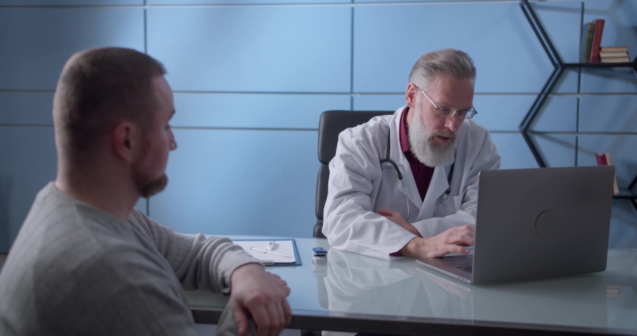 Professional serious focused middle aged hoary physician in medical coat sitting at table, consulting male patient about illness or surgery. Rear view man listening to old doctor at checkup meeting. Royalty-Free Stock Footage #1066951564