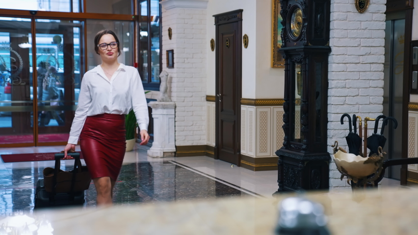 Charming lady with suitcase in the reception lobby. Girl approaching hotel reception to take the key. Portrait of gorgeous woman pressing bell button at reception desk.