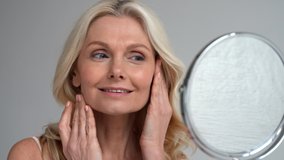 Happy 50s middle aged woman model touching face skin looking in mirror. Smiling mature older lady pampering, enjoying healthy skin care, aging beauty, skincare treatment cosmetic products concept. - Powered by Shutterstock - Get 15% off with code: PIKWIZARD15