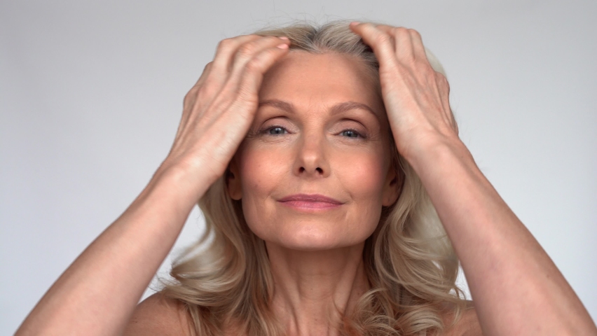 Happy 50s middle aged mature woman touching facial skin looking at camera pampering in mirror. Old healthy dry skin care beauty concept, skincare treatment, cosmetics and anti age plastic surgery. Royalty-Free Stock Footage #1066964725