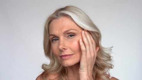Happy 50s middle aged mature woman touching facial skin looking at camera pampering in mirror. Old healthy dry skin care beauty concept, skincare treatment, cosmetics and anti age plastic surgery.