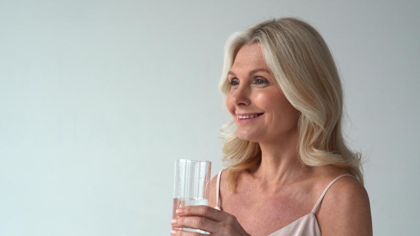 Smiling healthy thirsty fit mid aged 50s woman holding glass drinking water. Happy mature lady hydrating body with clean filtered fresh mineral water for skin care, beauty, diet nutrition hydration. Royalty-Free Stock Footage #1066964737