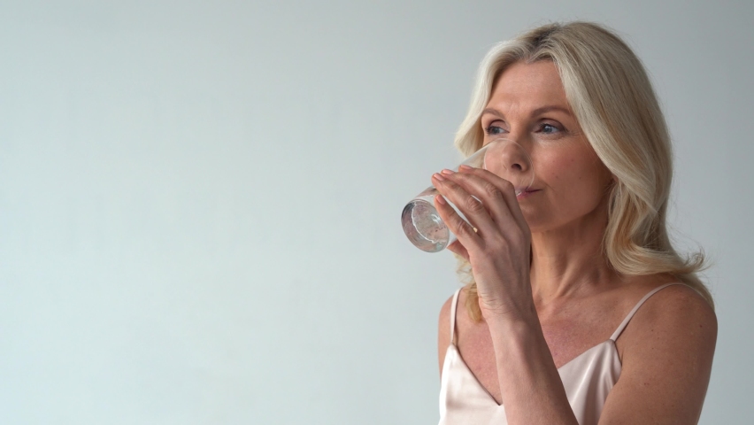 Smiling healthy thirsty fit mid aged 50s woman holding glass drinking water. Happy mature lady hydrating body with clean filtered fresh mineral water for skin care, beauty, diet nutrition hydration. Royalty-Free Stock Footage #1066964737
