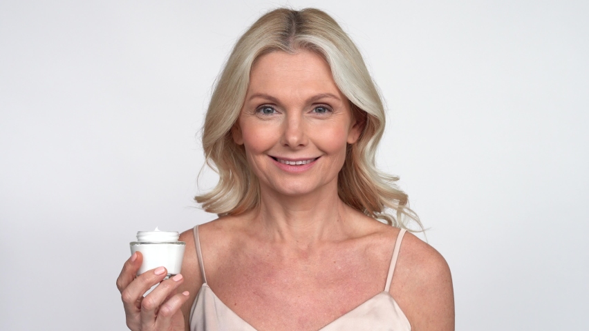 Smiling 50s middle aged mature woman putting tightening facial cream on face looking at camera. Anti age healthy dry skin care beauty therapy concept, skincare rejuvenation treatment against wrinkles. Royalty-Free Stock Footage #1066964740