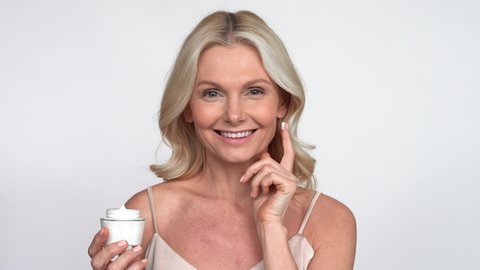 Smiling 50s middle aged mature woman putting tightening facial cream on face looking at camera. Anti age healthy dry skin care beauty therapy concept, skincare rejuvenation treatment against wrinkles.