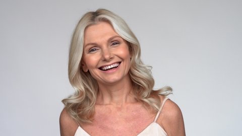 Happy smiling beautiful 50s middle aged mature woman looking at camera, laughing on white background. Anti age face beauty, skin and body care, wellness and self care concept. Close up portrait.