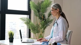 A friendly female doctor in a medical uniform and eyeglasses consults a patient online, communicates on a video call using a laptop.  Online medical help