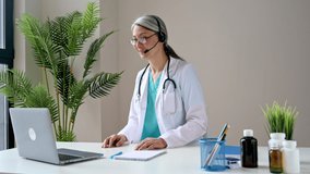 A friendly female doctor in a medical uniform and headphones consults a patient online, communicates on a video call using a laptop.  Online medical assistance