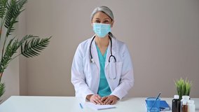 Middle aged gray-haired female doctor wears medical mask, looks at camera, consults the patient. Remote online medical consultation, medicine distance services, virtual medical help concept