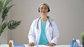 A friendly senior gray-haired female doctor wears headset looks at camera, conducts distance consultation. Remote online medical consultation, medicine distance services, virtual medical help concept