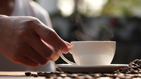 Men's hand takes hot coffee cup with coffee beans and natural steam smoke of coffee on wooden table, slow motion. Hot Coffee Drink Concept.