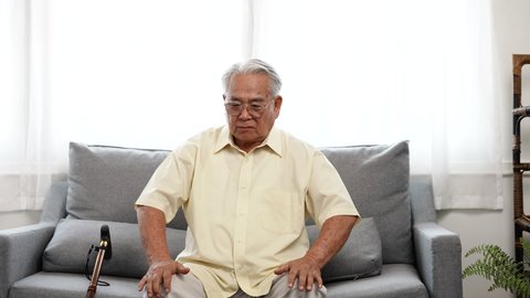 Asian Eldery senior man feeling pain in knee and standing up step walking the floor with a cane at home in living room alone. Learn how to be alone after retirement concept.